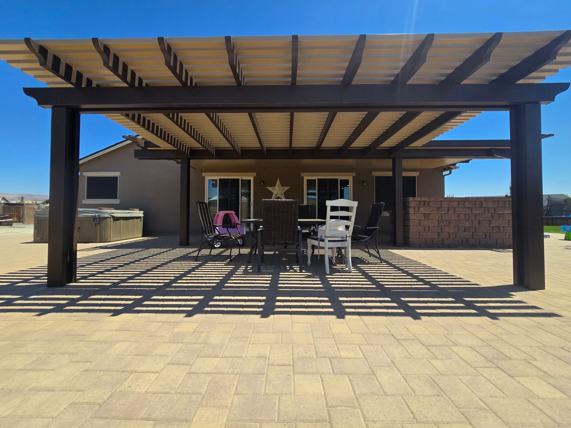 Pergola over patio with furniture, casting shadows on the paved ground under a bright blue sky.