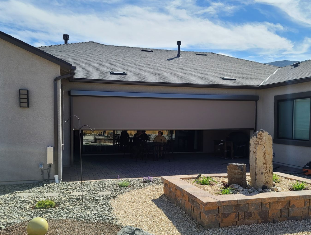 A tan retractable shade partially covers a patio, revealing people inside. The patio has a rock garden and brick edging.