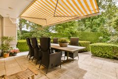 Patio with dining table and chairs shaded by yellow-striped awning; greenery in the background.