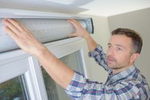 Man installing or adjusting a white roller blind over a window.