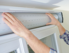 Hands installing a white roller shutter above a window frame.