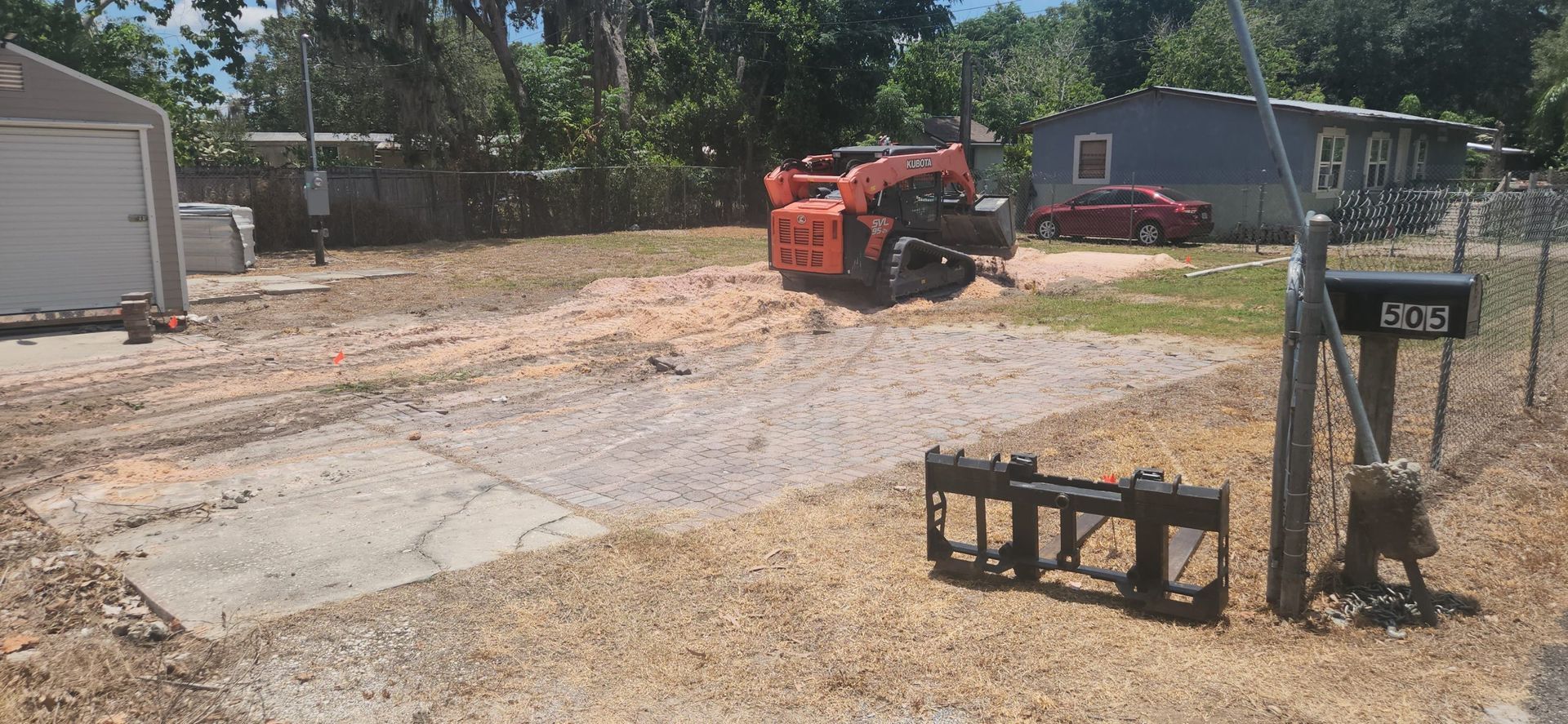 An orange skid steer operating on a cleared lot. A house and garage are visible in the background, with a red car parked in front of the house.