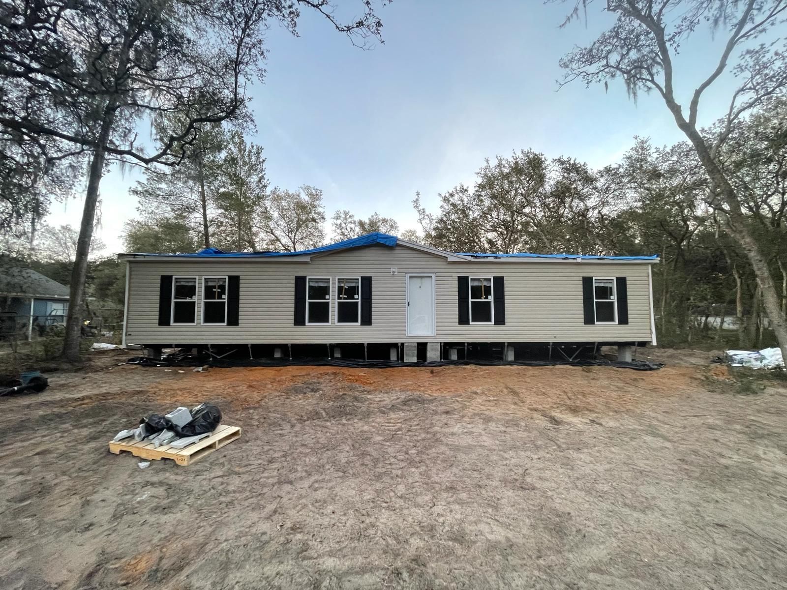 Mobile home on blocks with blue tarp covering part of the roof, surrounded by trees on a dirt lot.