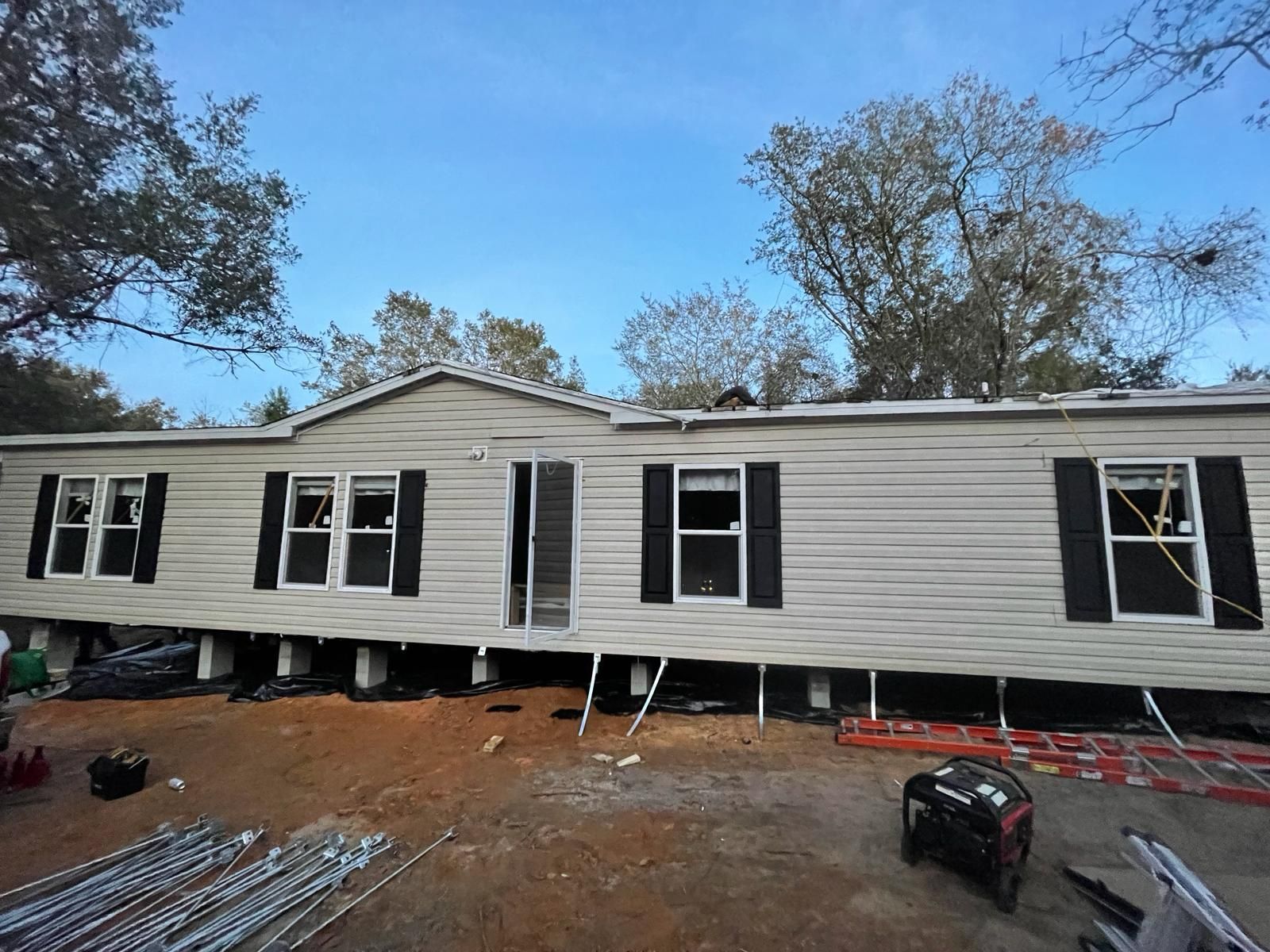 A light-colored mobile home with black shutters, undergoing renovation. The home sits on pillars with construction materials nearby under a blue sky.