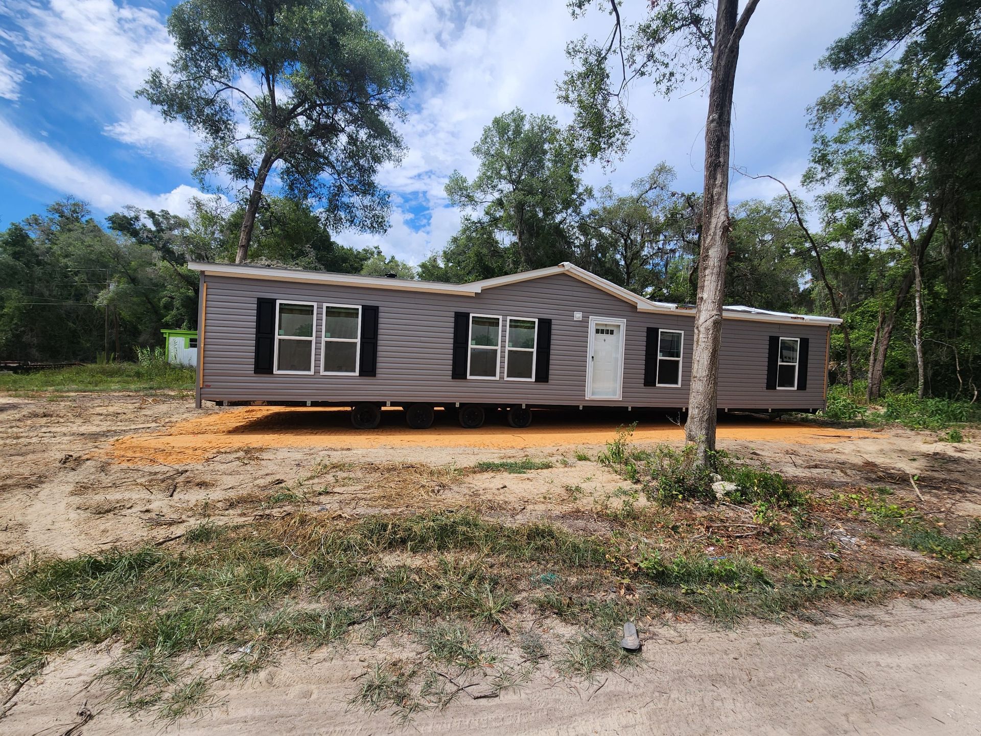 A single-story, gray mobile home with black shutters sits on a sandy lot, surrounded by trees under a partly cloudy sky.