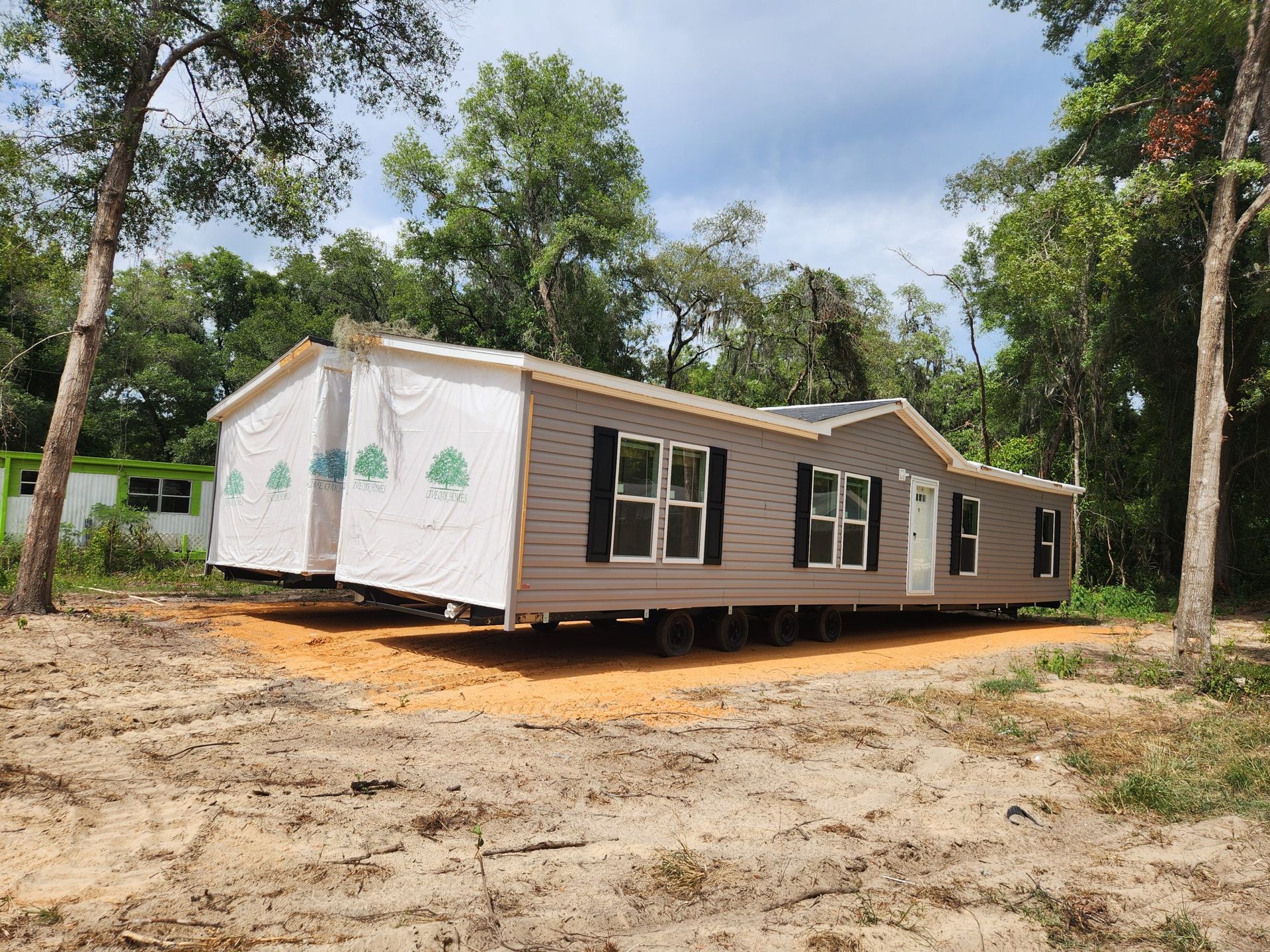 A tan and brown modular home sits on a gravel pad in a wooded area. The home has black shutters and white trim.