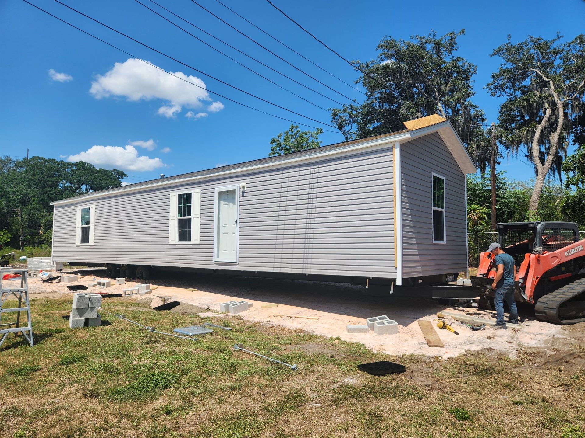 A mobile home is being set onto concrete blocks in a grassy area. A construction worker operates an orange skid steer nearby.