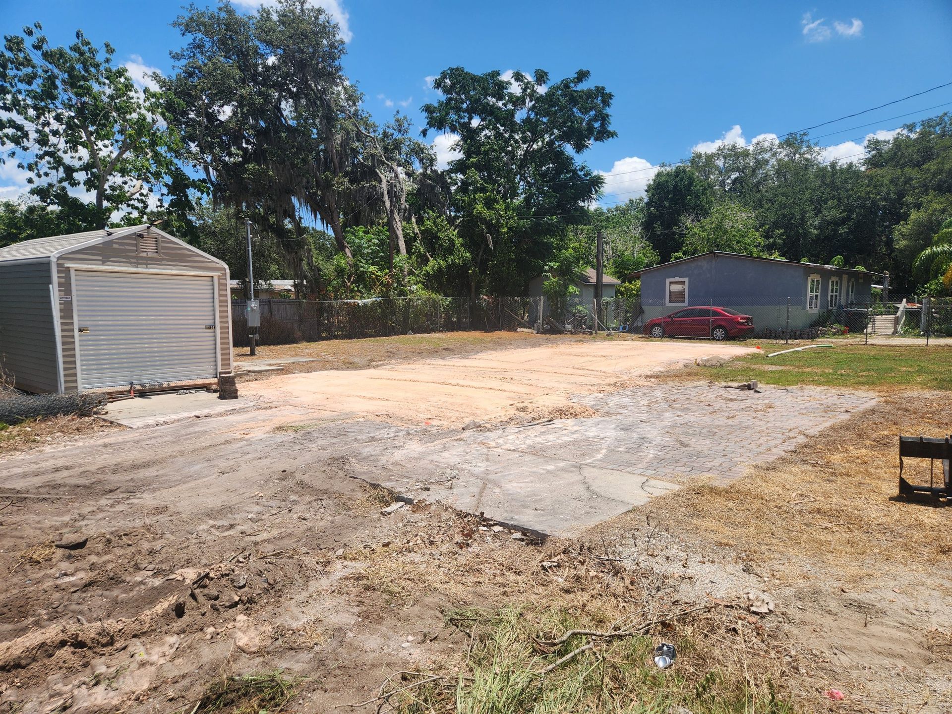 An empty lot with a concrete pad and dirt, garage on the left, and a blue house in the background. Bright sunny day.