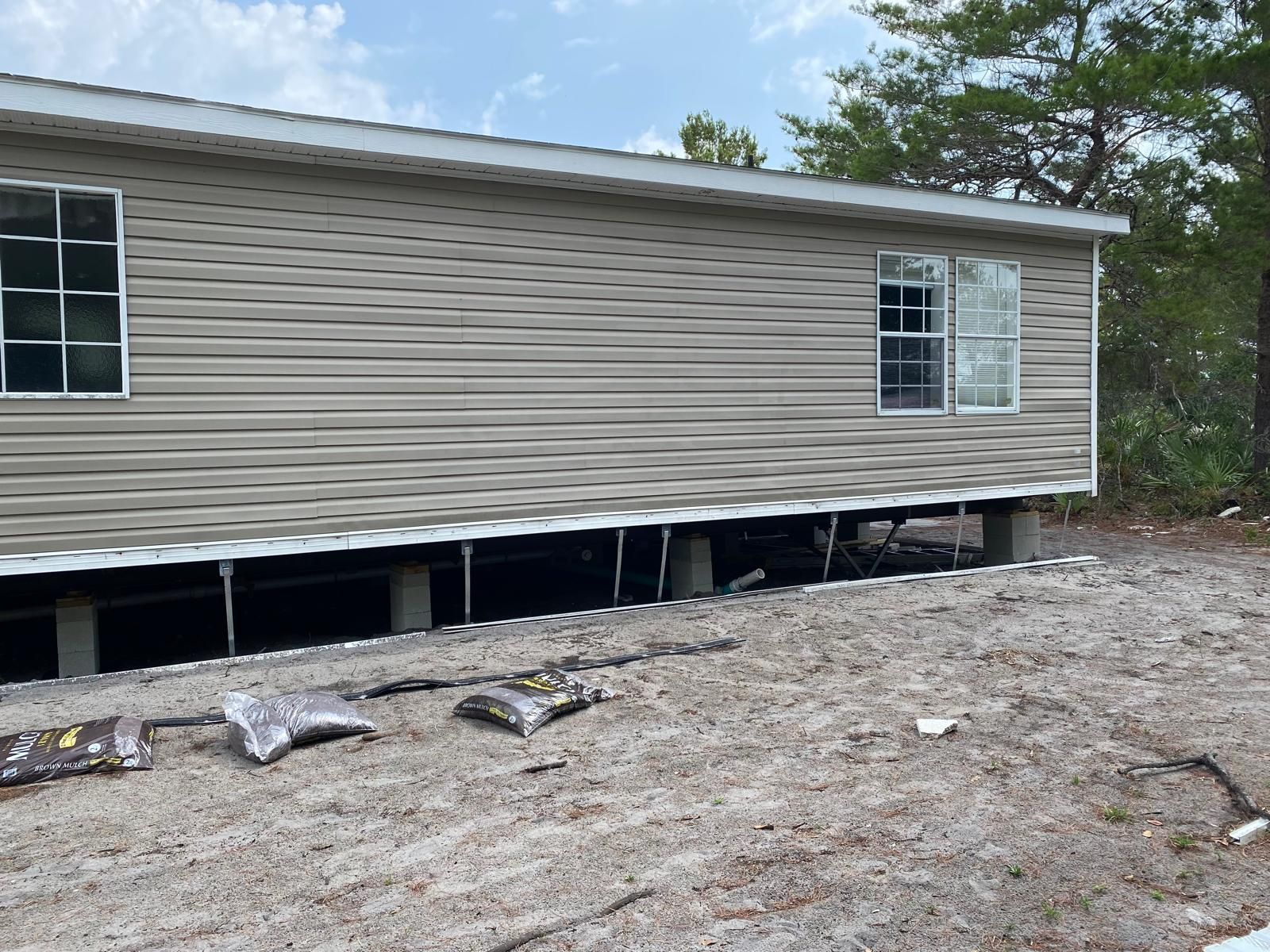 Tan mobile home on concrete supports, sitting on sandy ground. Windows visible, trees in background.