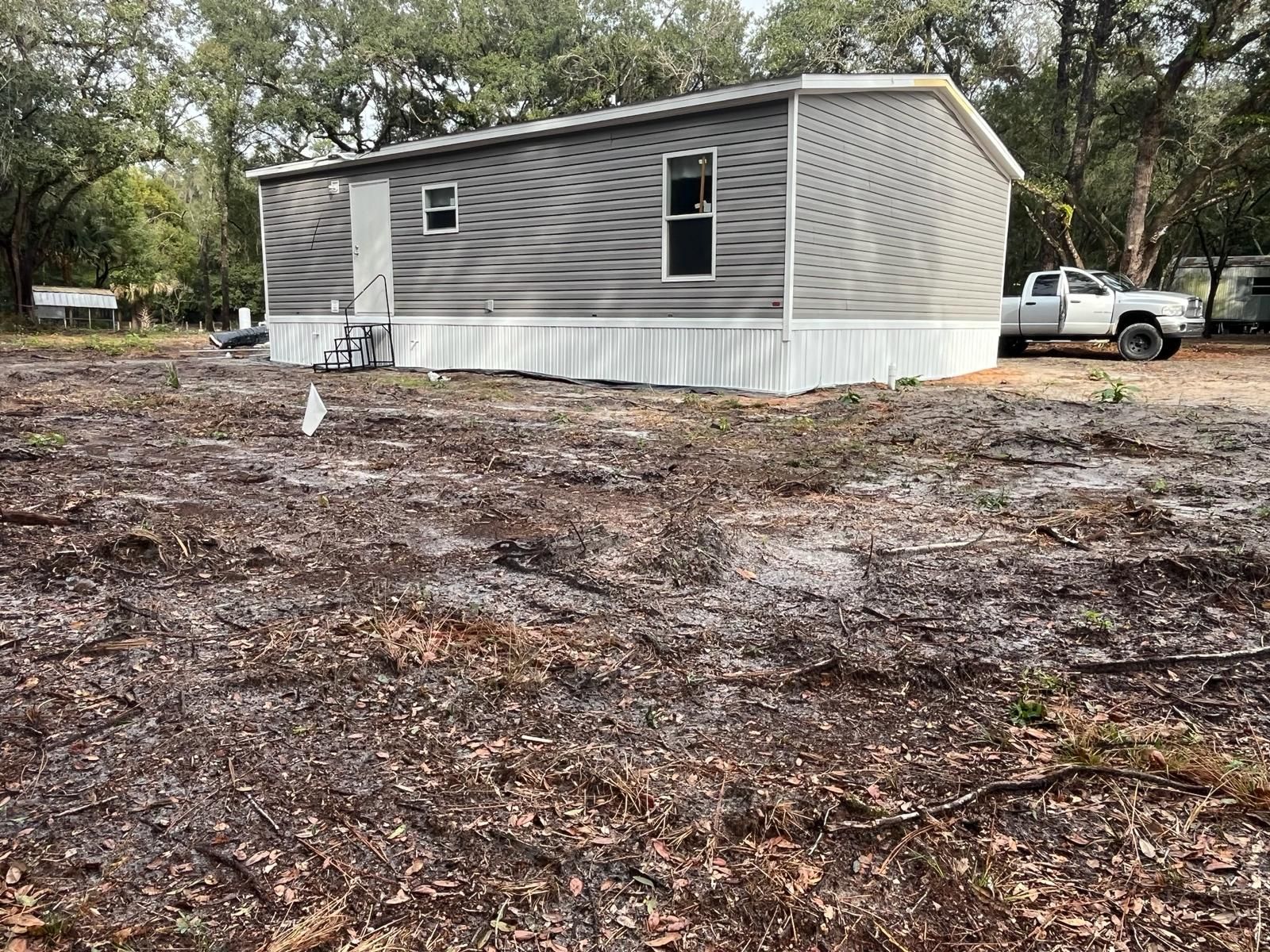 Mobile home with gray siding and white trim sits on a sandy lot, with a truck parked nearby.