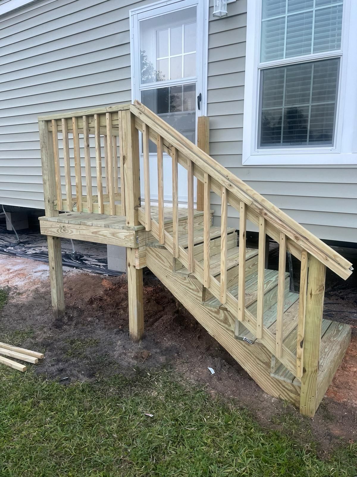 Wooden exterior staircase with railings and a small landing leading to a house entrance with two windows. Light green siding and grass are visible.