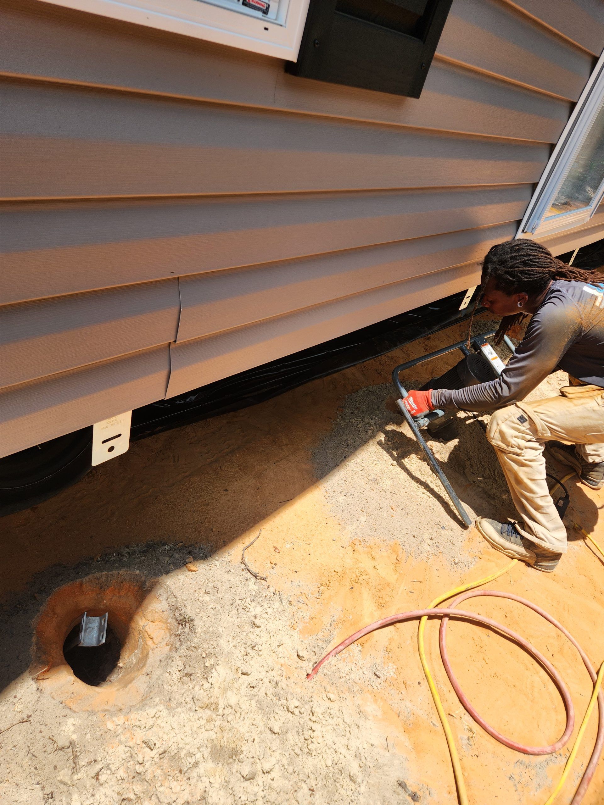 A person works on the side of a building, near a concrete surface with exposed pipes. They appear to be cutting siding.