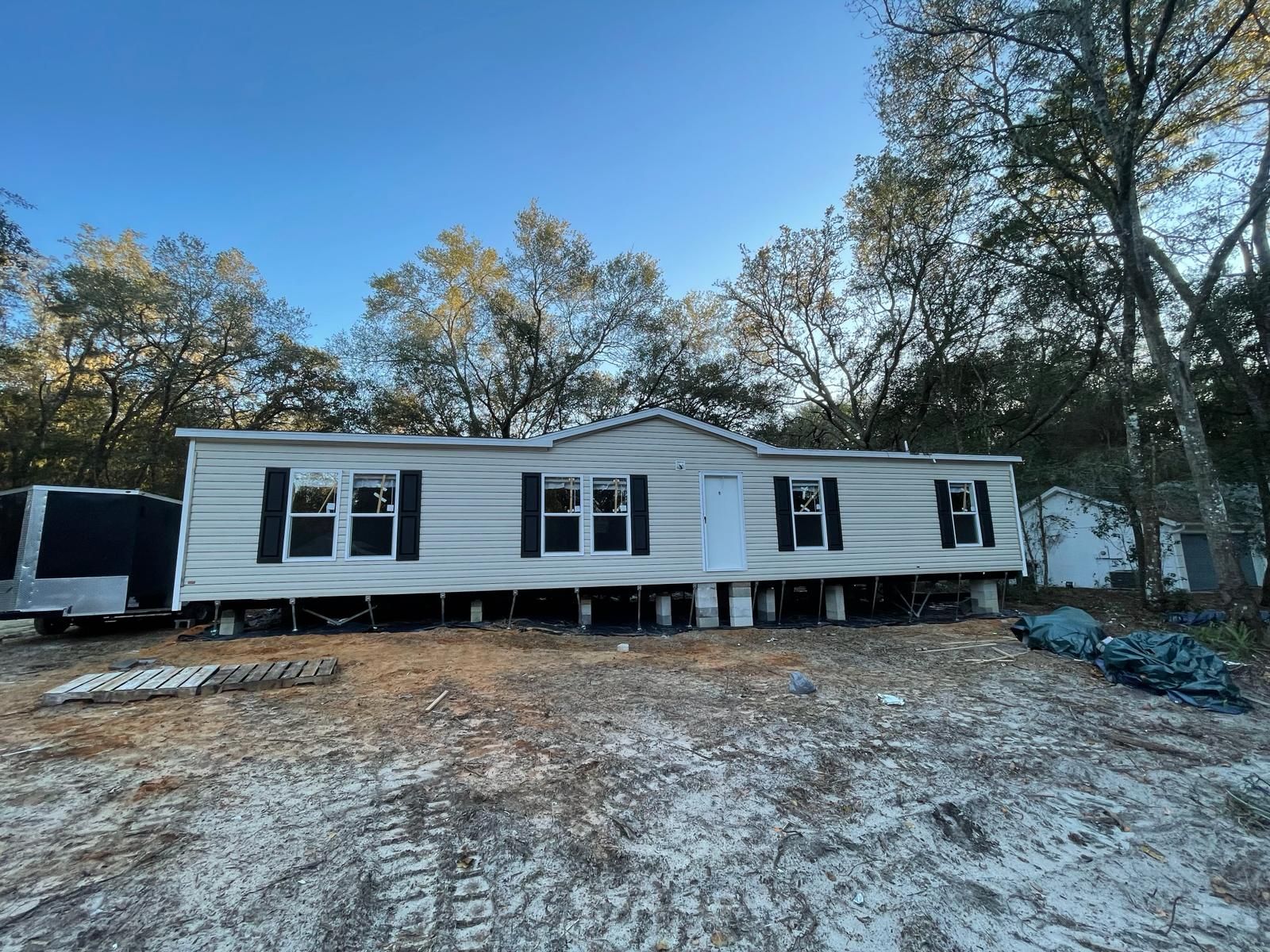 A single-wide mobile home on concrete blocks in a sandy area with trees in the background under a blue sky.