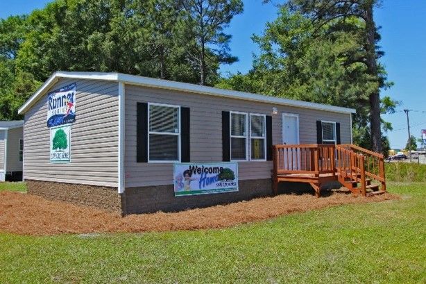 A light brown mobile home with black shutters and a small wooden deck sits on a green lawn, under a blue sky.