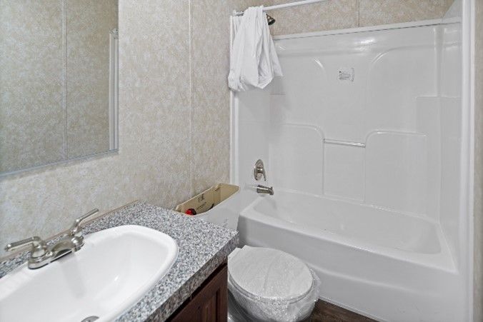 Bathroom with a white bathtub, toilet, and sink. Beige walls and a gray countertop with dark brown cabinets. A white towel hangs on the shower rod.