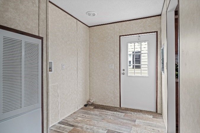 An entryway in a home with a white door, light-colored walls, and wood-look flooring. A large white vent cover is visible on the left.