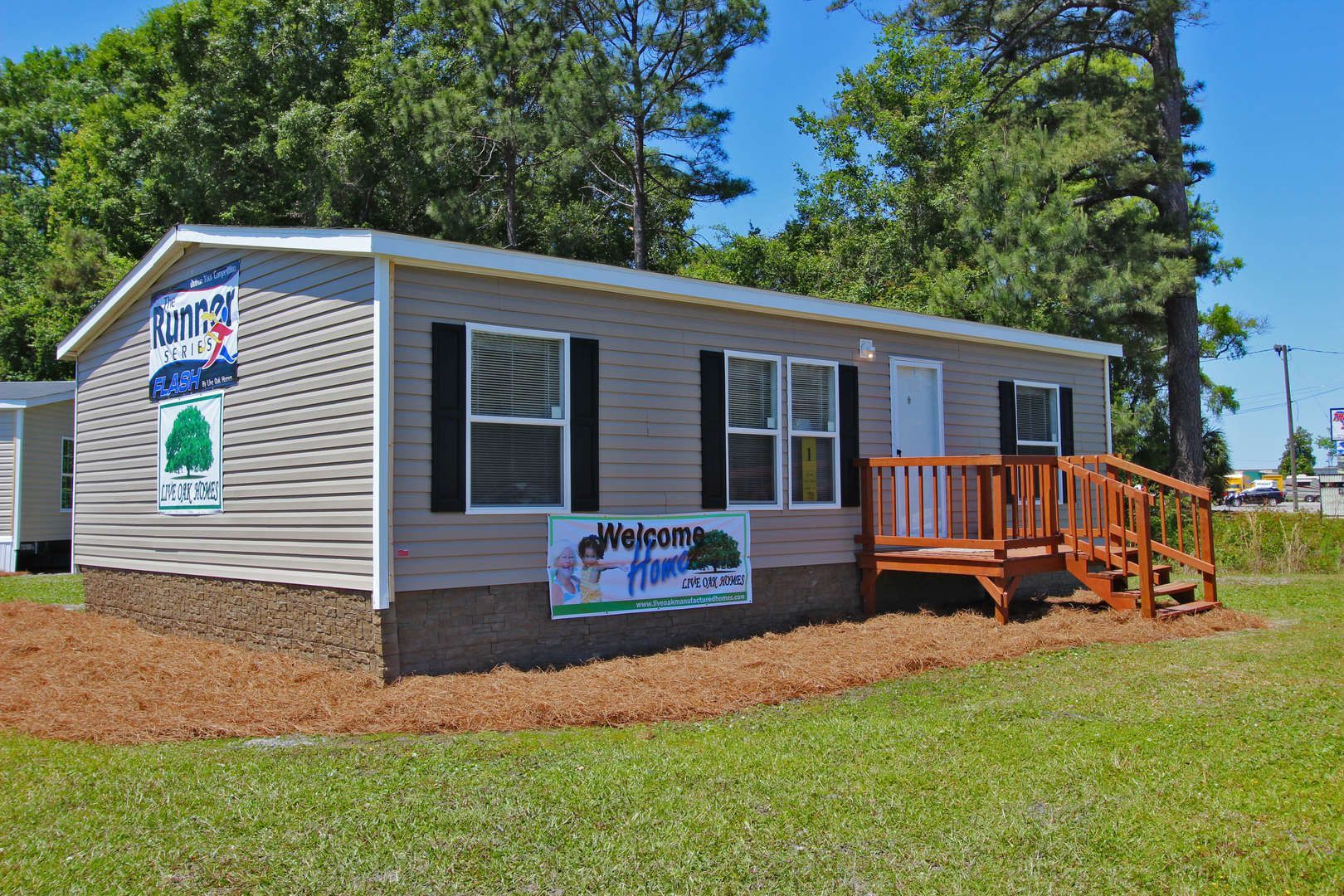A light grey manufactured home with brown trim and a small wooden porch, on a grassy lawn.