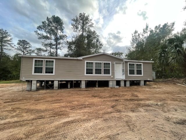 Tan mobile home elevated on concrete blocks, set in a dirt clearing with trees in the background, under a cloudy sky.