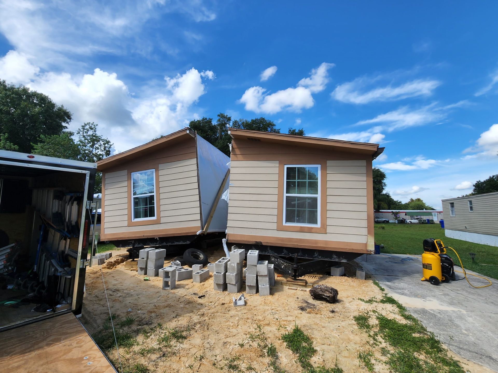 Two tan mobile home sections being installed on concrete blocks under a blue sky.