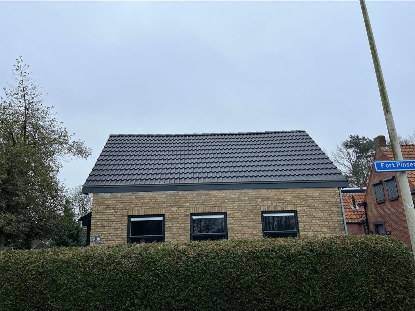 een bakstenen huis met een pannendak en een heg ervoor.