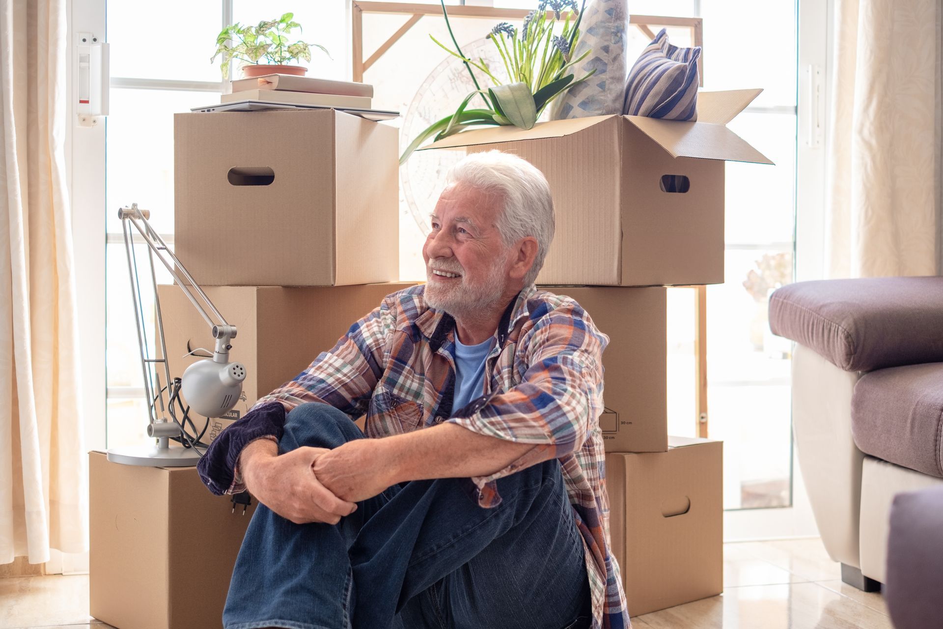 Older man sits near moving boxes, smiling. Sunlight streams through window in a bright living room.