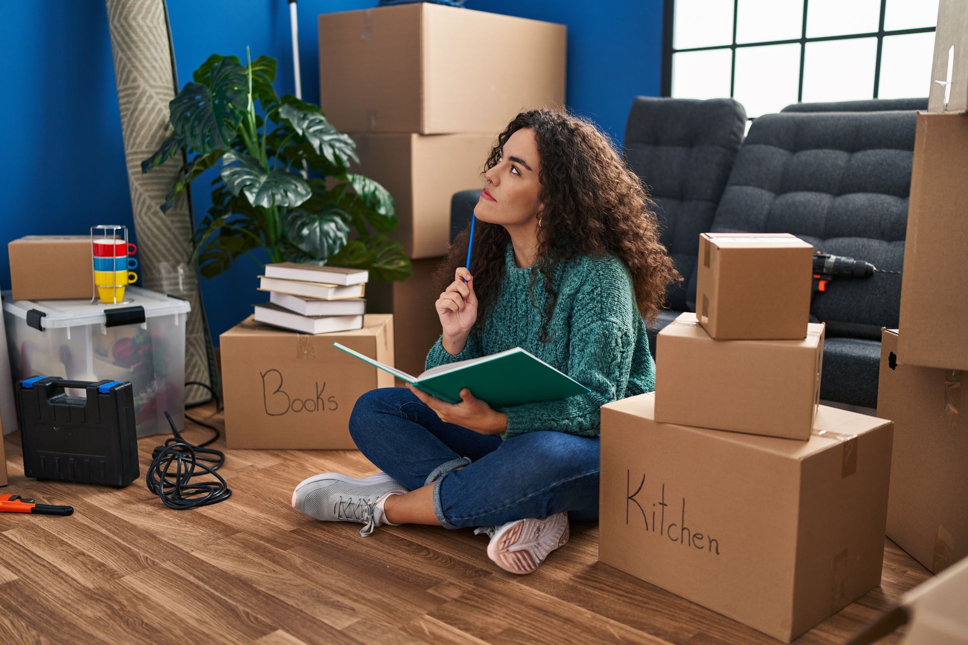 Woman sits on the floor cross-legged among moving boxes, writing in a notebook, looking thoughtful.