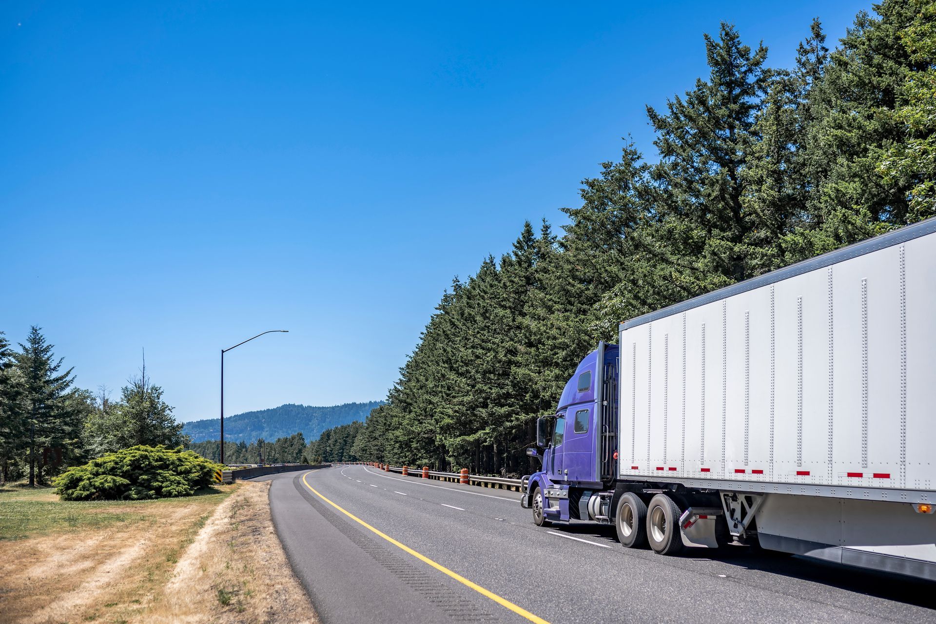 A purple semi truck is driving down a highway next to trees.