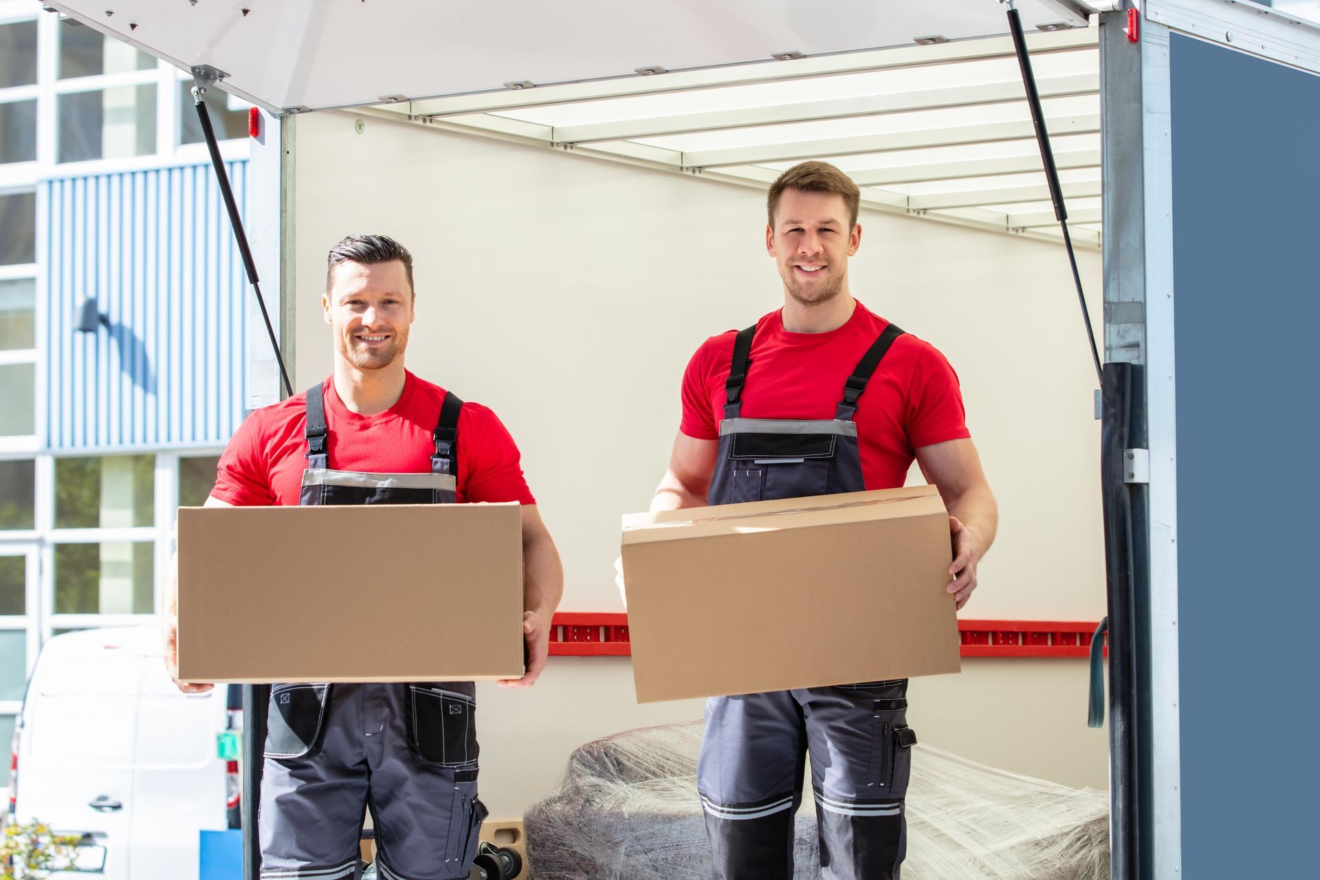 Two smiling movers in red shirts and gray overalls, holding boxes at the back of a truck.
