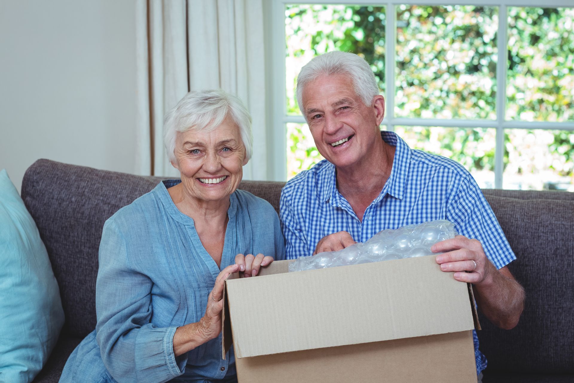 Senior couple smiles while opening a cardboard box on a couch. Window in background.