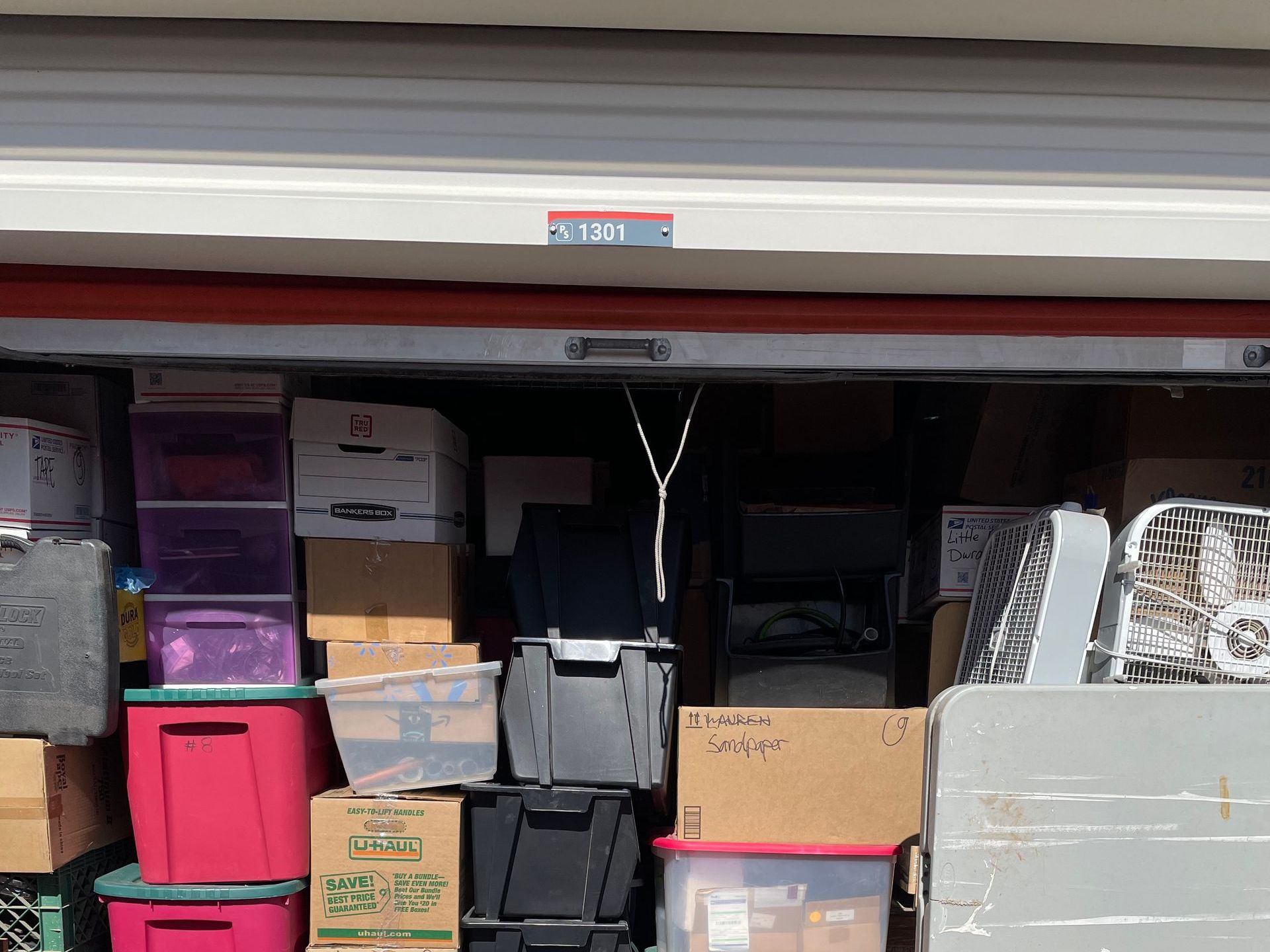 Storage unit filled with boxes, bins, and various items, including a fan. The unit door is open.