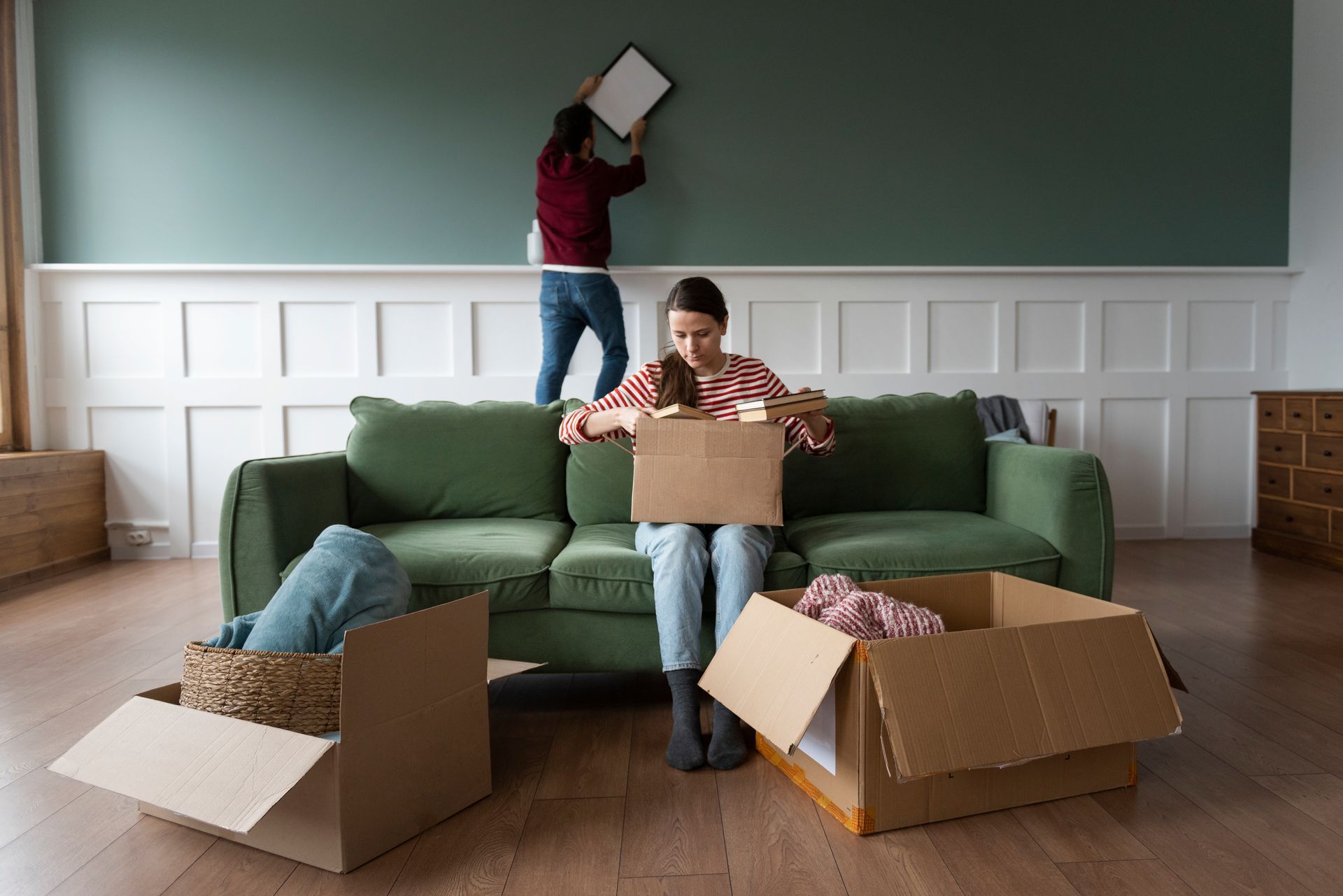 Woman unpacking boxes, man hanging a picture on wall in a living room with green sofa and white paneling.