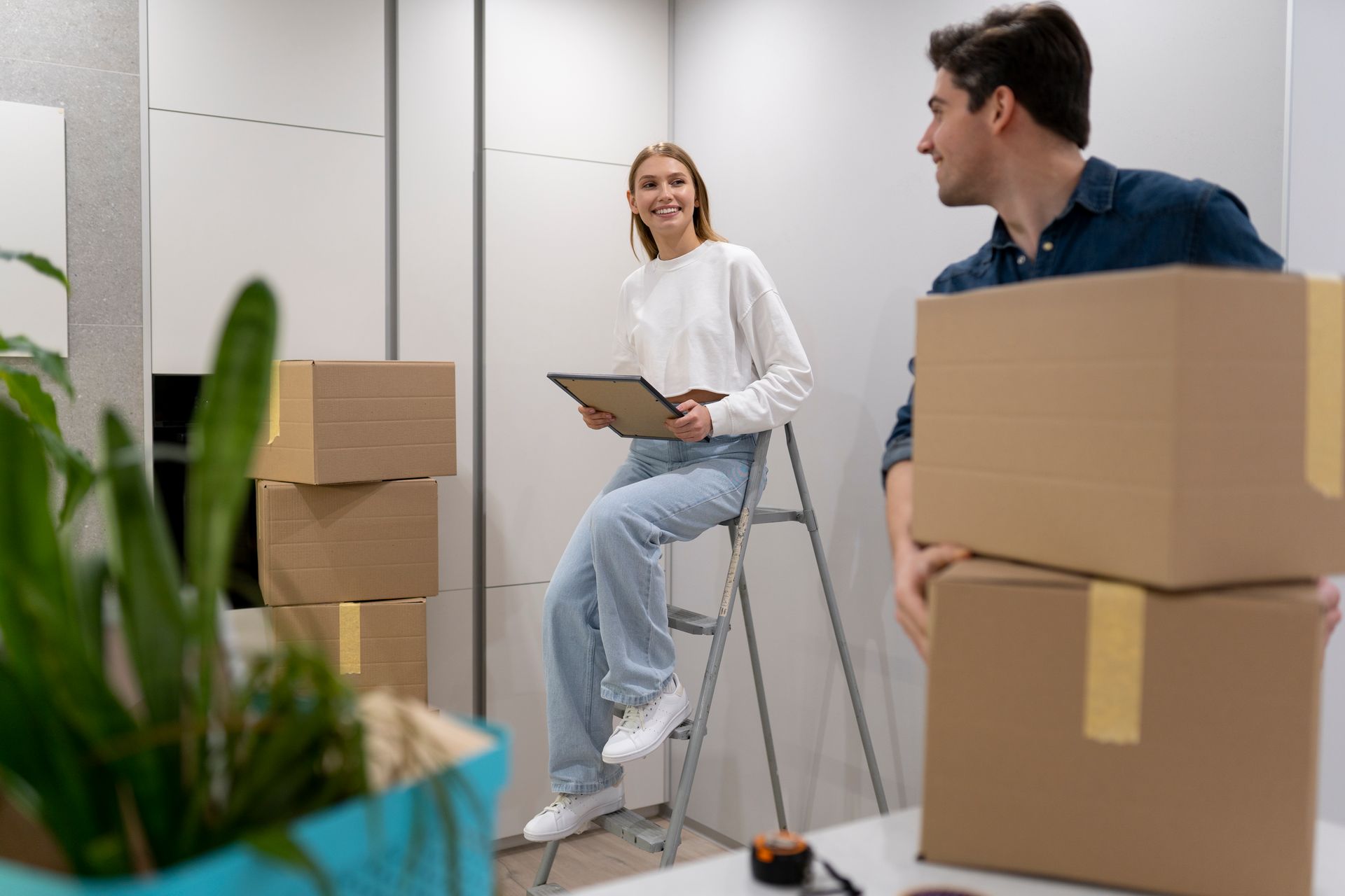 Couple unpacking, one on stepladder holding clipboard, surrounded by moving boxes.
