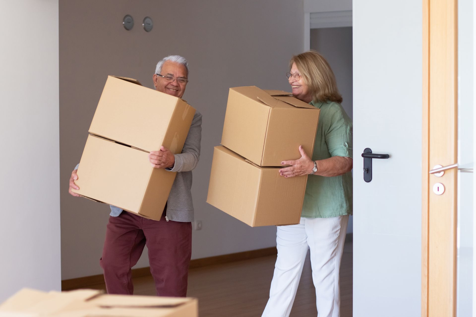 Elderly couple moving boxes into a new home, smiling near an open door.