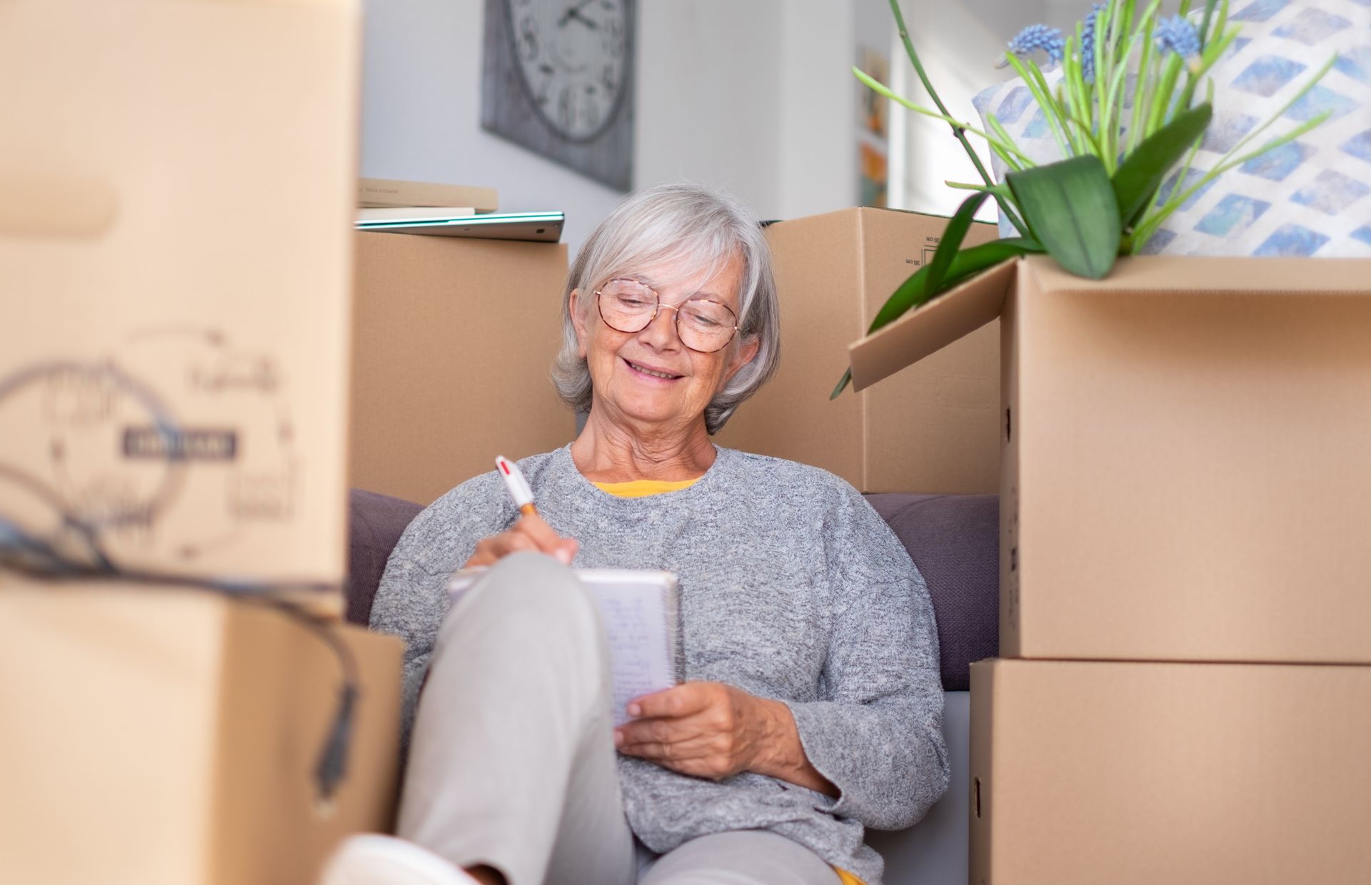 Woman with glasses sits among boxes, smiling while writing in a notebook.