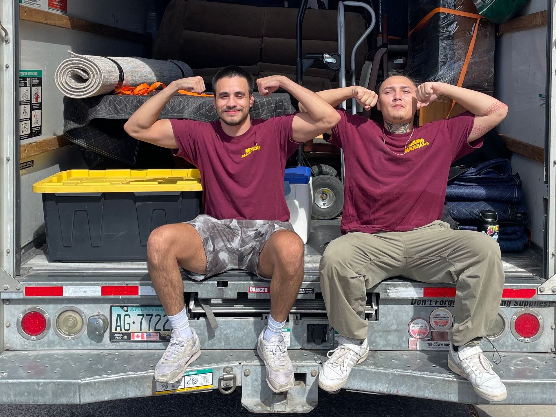 Two men are sitting on the back of a moving truck.