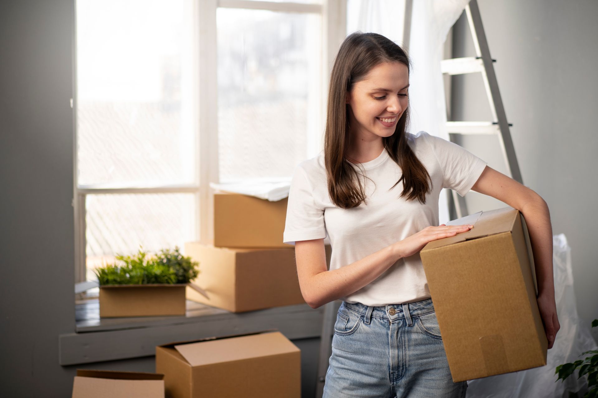 Woman in a white t-shirt and jeans smiles while holding a moving box near a window with more boxes.