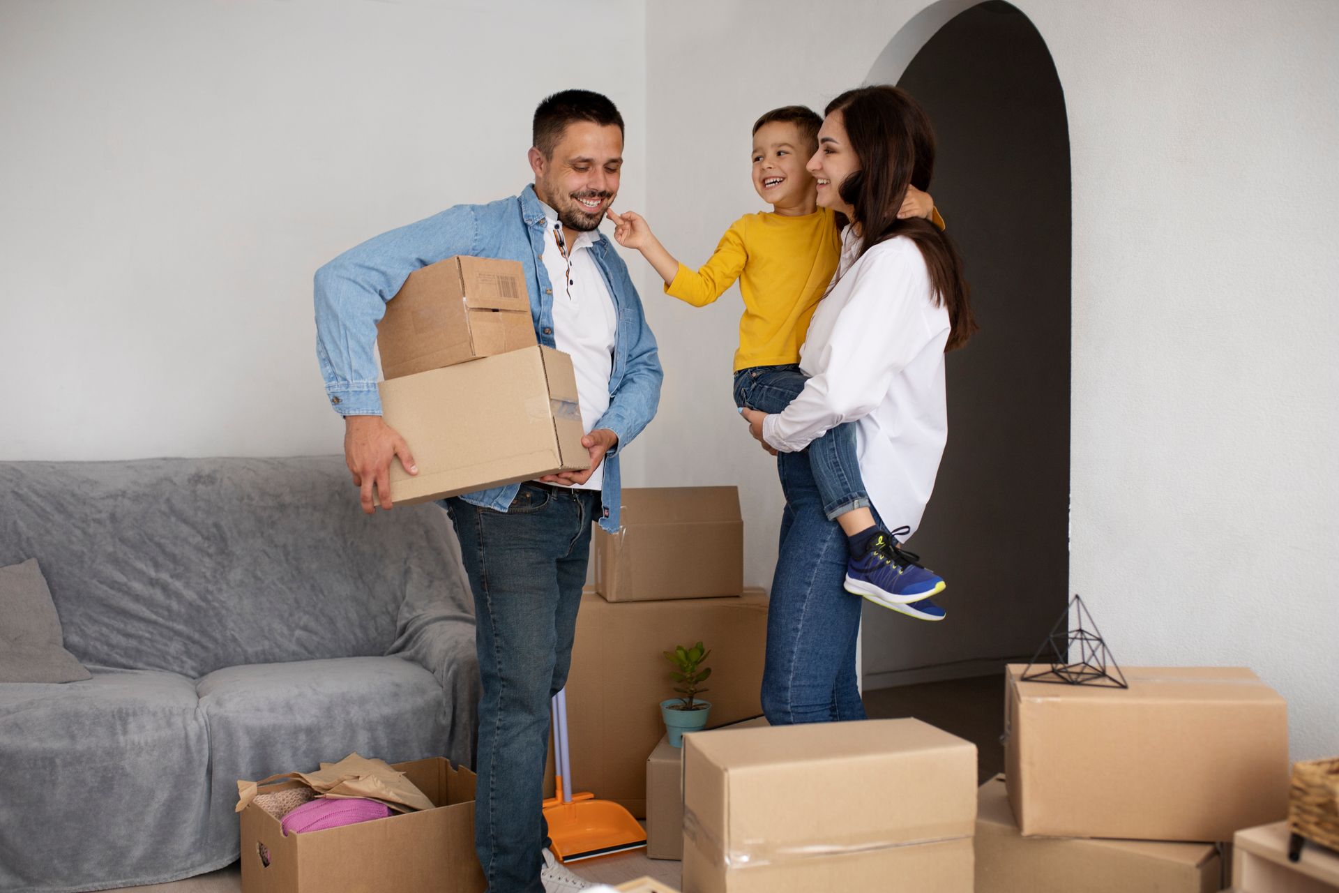 Family in new home, surrounded by boxes. Father holding boxes, mother holding child, smiling.