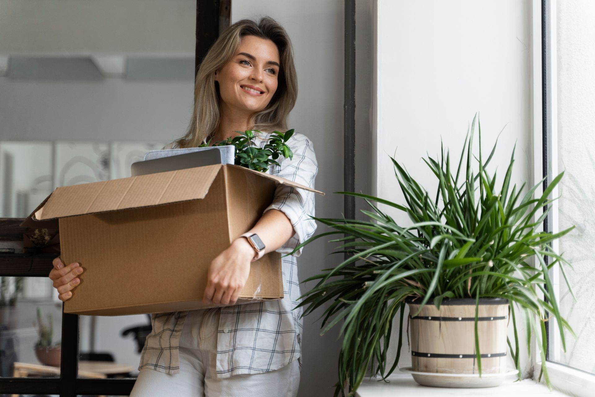 Woman holding a moving box smiles near a potted plant and window.