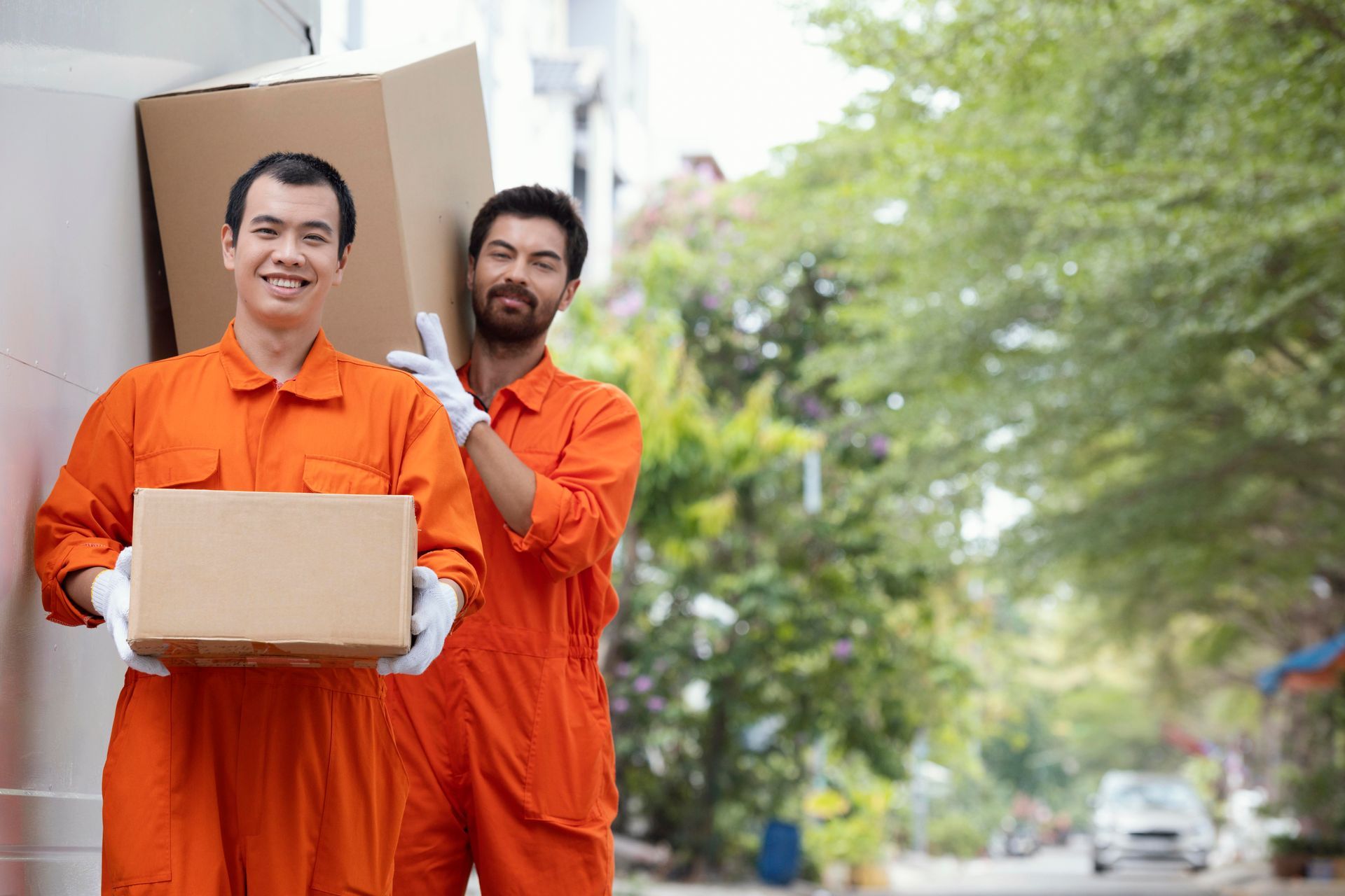 Two movers in orange jumpsuits carrying cardboard boxes outdoors; one smiling.