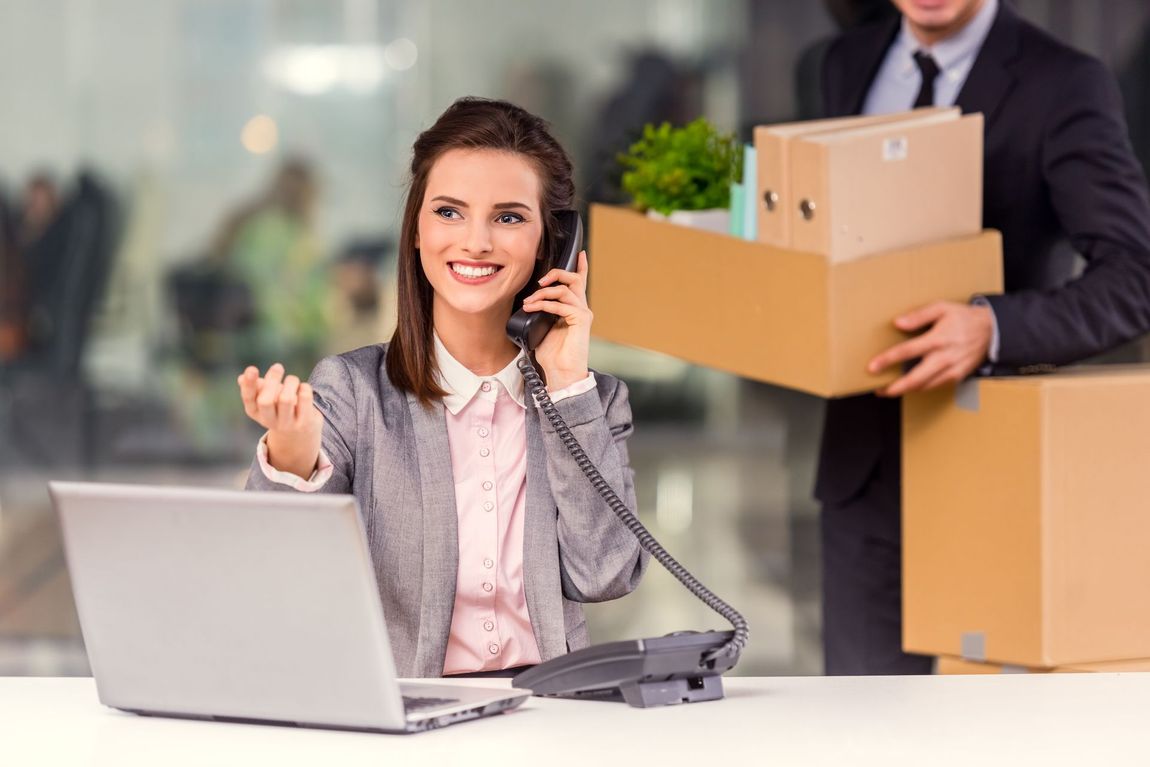 A woman is sitting at a desk talking on a phone while a man is carrying boxes.