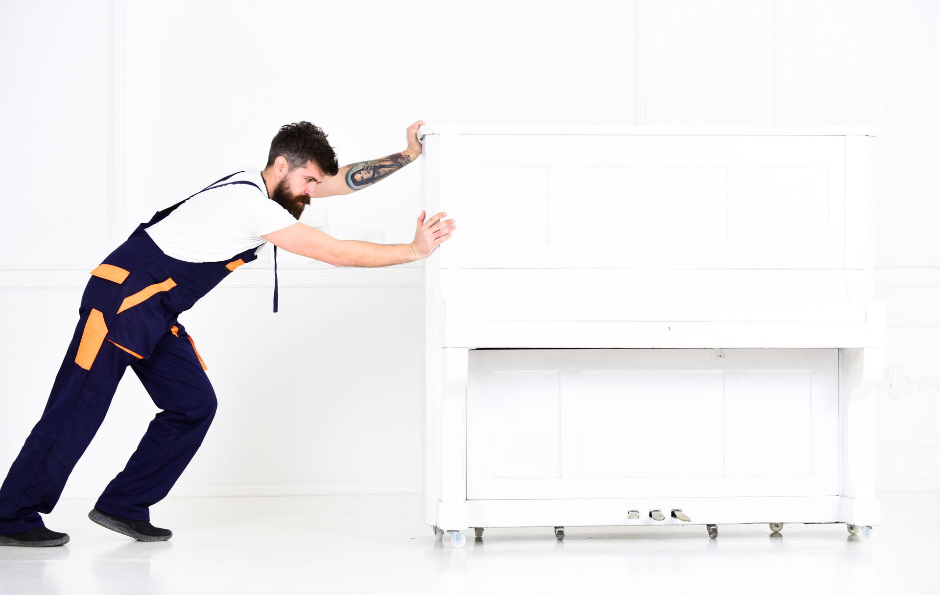 Man in blue overalls pushing a white piano in a white room.