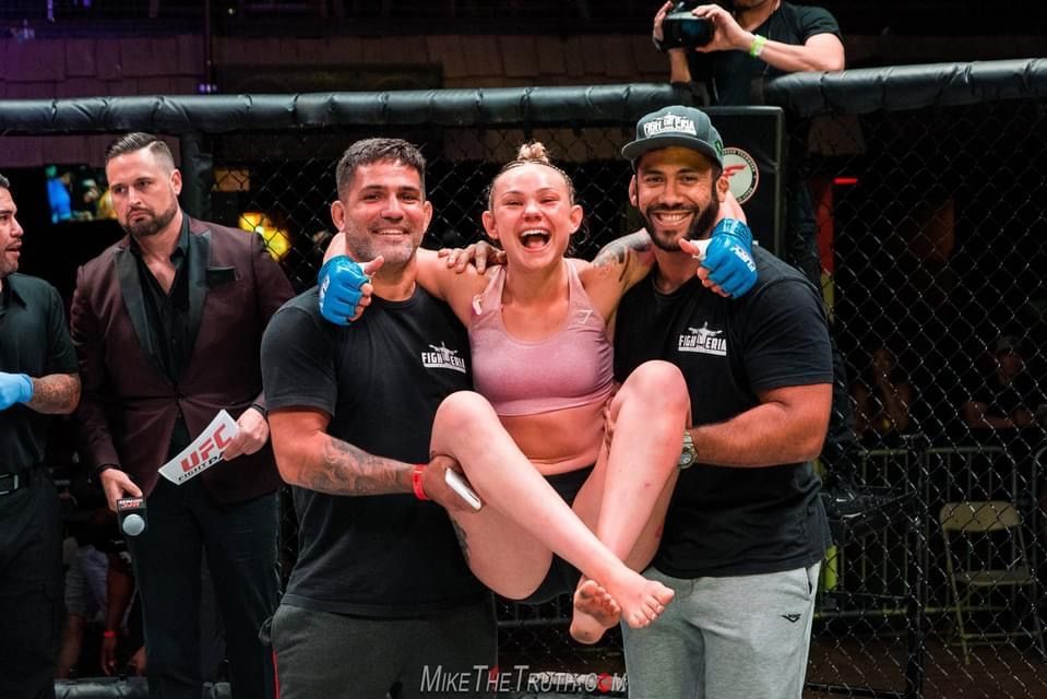 Woman held up by two men in MMA cage, smiling with excitement.