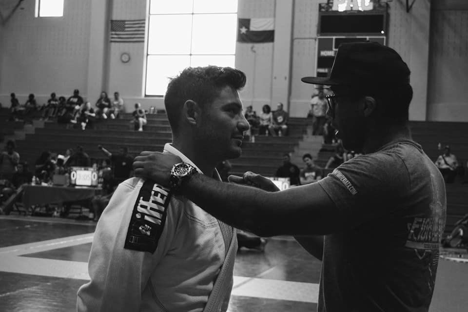 Man in a white gi receives a medal from another person in a gymnasium, with spectators in the background.