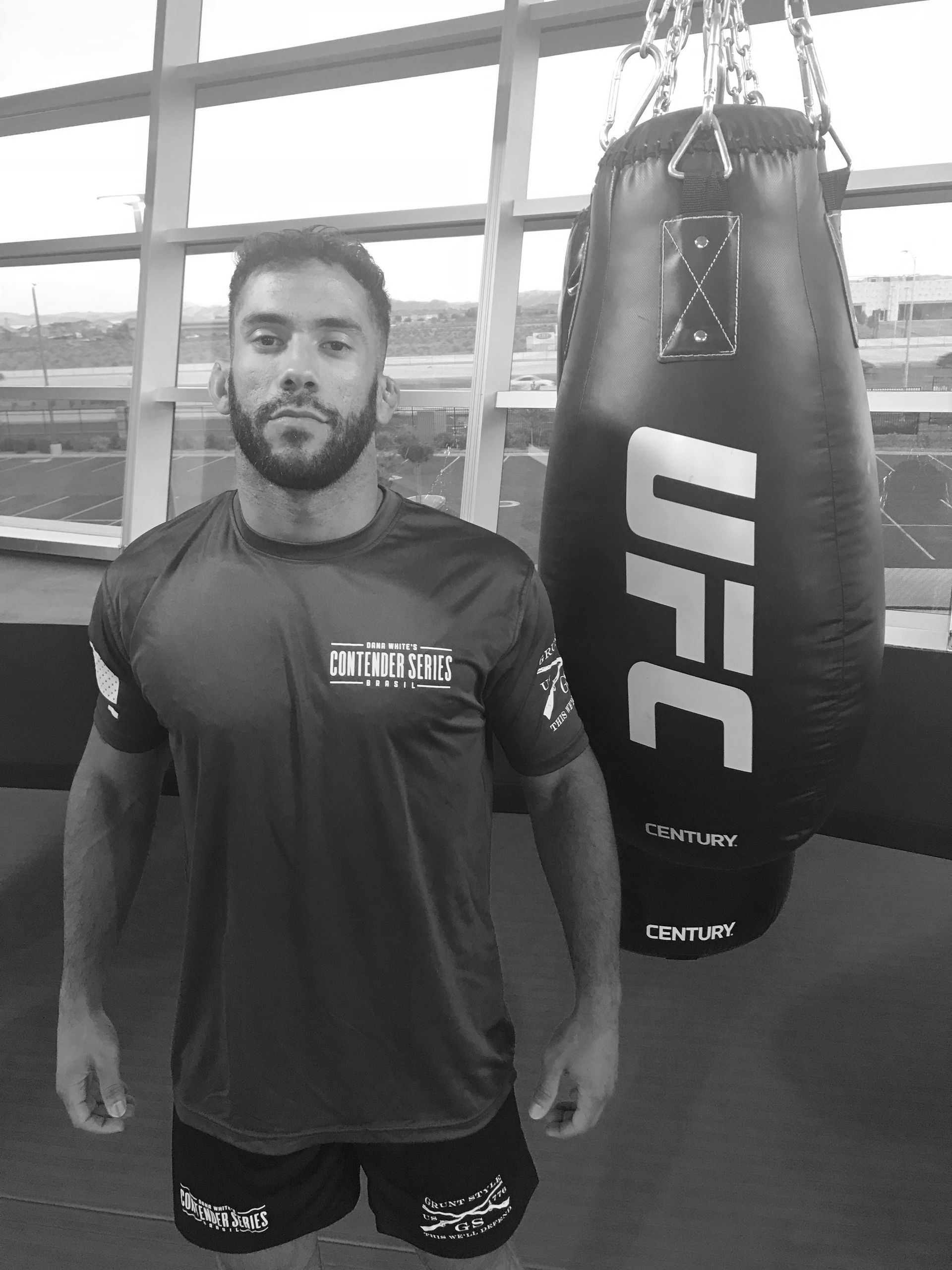 Man standing next to a UFC punching bag in a gym. He wears a shirt and shorts.