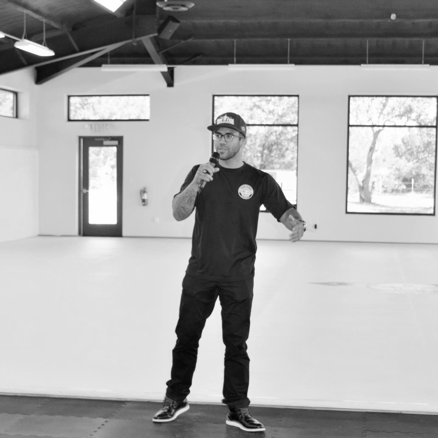 A person in a t-shirt and cap speaks into a microphone while standing in a large, empty, light-filled studio.