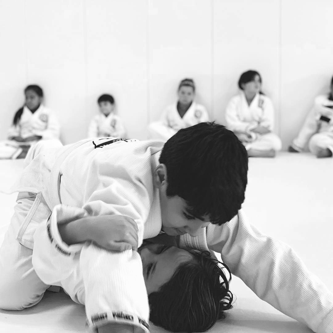 Two children in white martial arts uniforms practicing a ground grappling technique on a mat, with classmates observing.