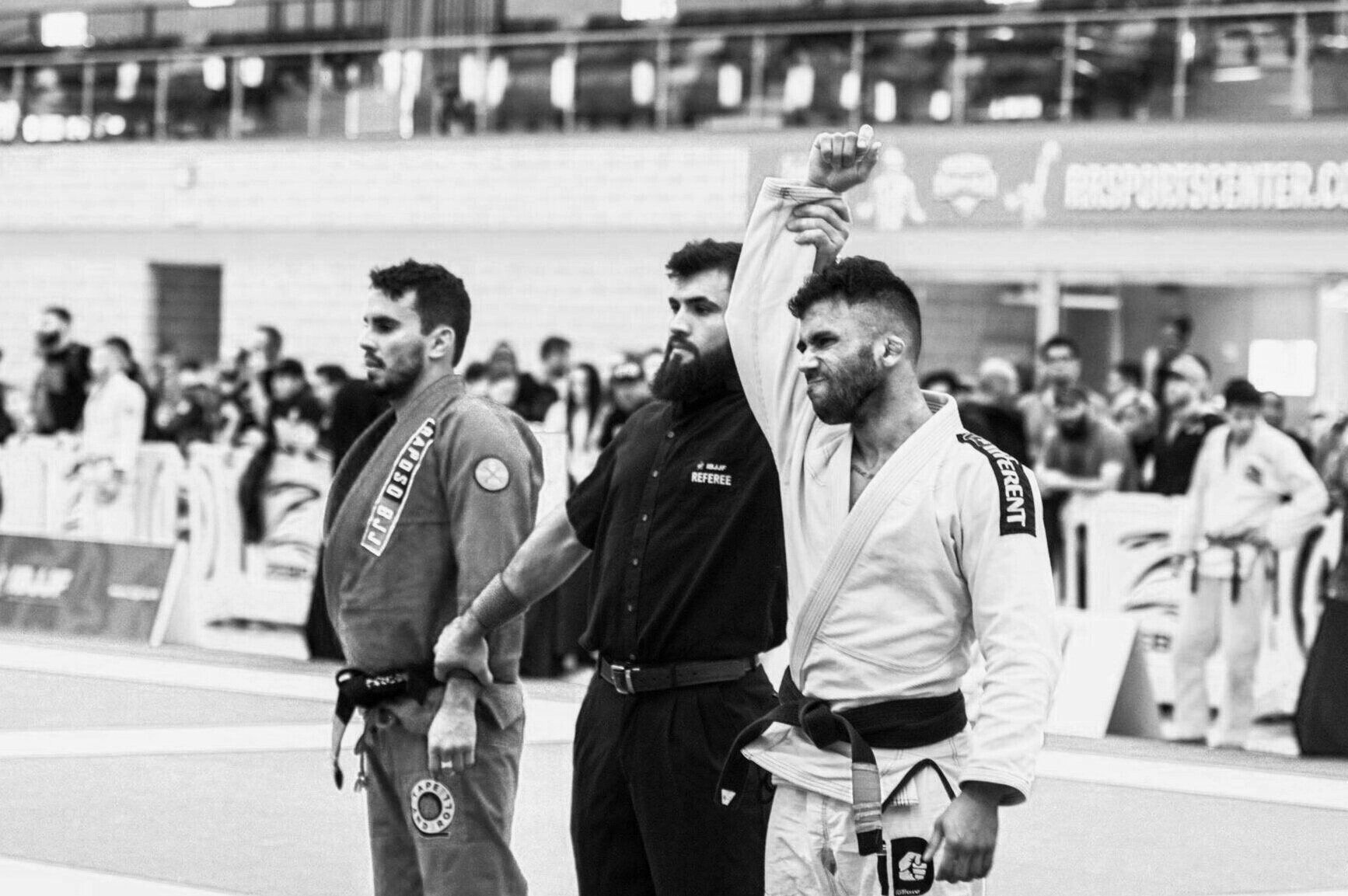 A referee raises the arm of a man in a gi, declaring him the winner of a jiu-jitsu match.