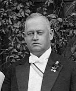 Ein Mann im Smoking und mit Fliege steht vor einem Busch.