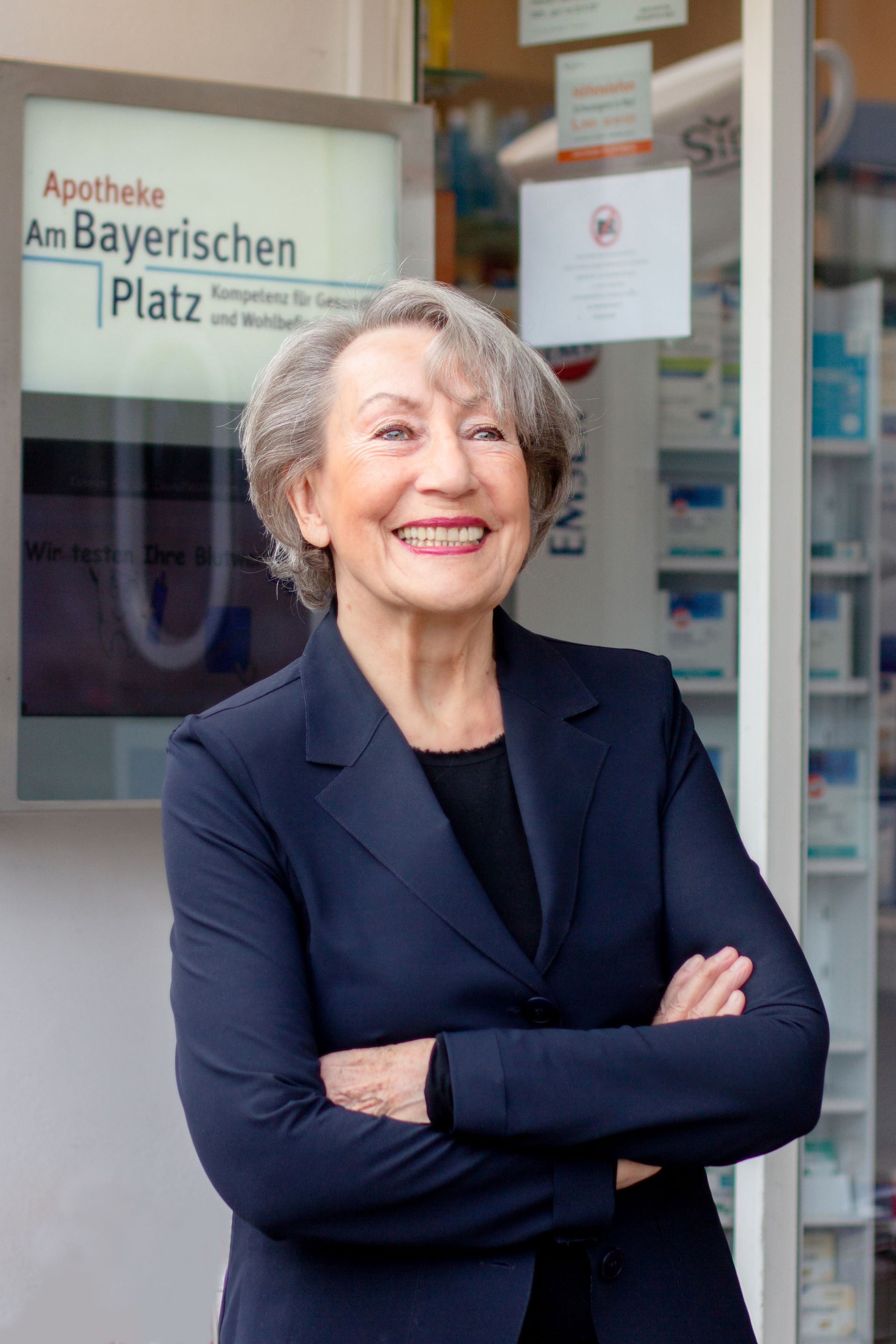 Eine Frau in einem blauen Anzug steht mit verschränkten Armen vor einer Apotheke und lächelt.