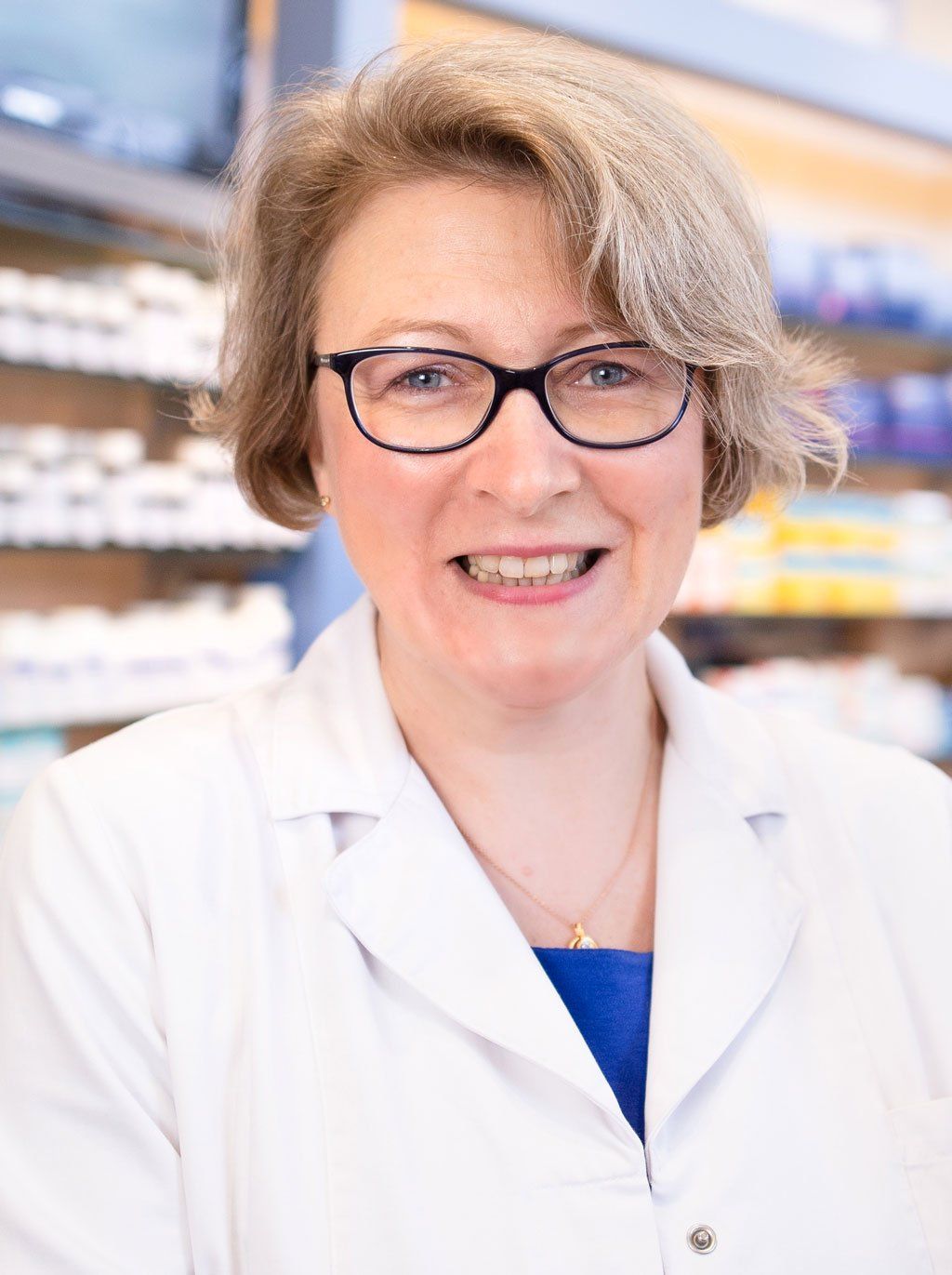 Eine Frau mit Brille und weißem Kittel lächelt in einer Apotheke.